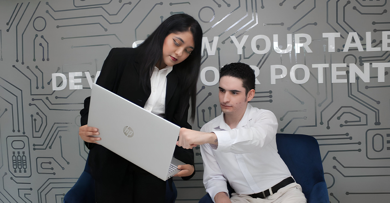 "Two young professionals collaborating in a modern tech office; a woman stands holding an HP laptop while a male colleague sits in a blue chair, pointing at the screen against a wall with circuit board graphics and the partial text 'GROW YOUR TALENT'."