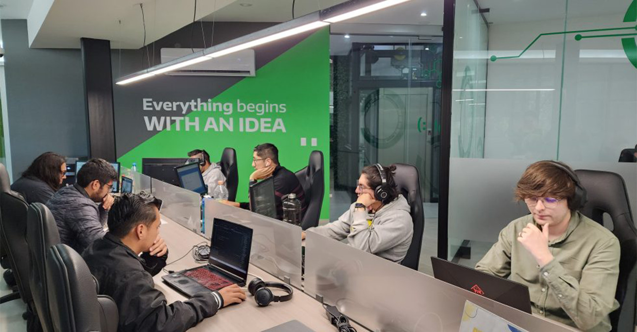 "A group of IT professionals working on laptops and computers at a long shared desk in a modern office, with a green wall in the background that reads 'Everything begins WITH AN IDEA'."